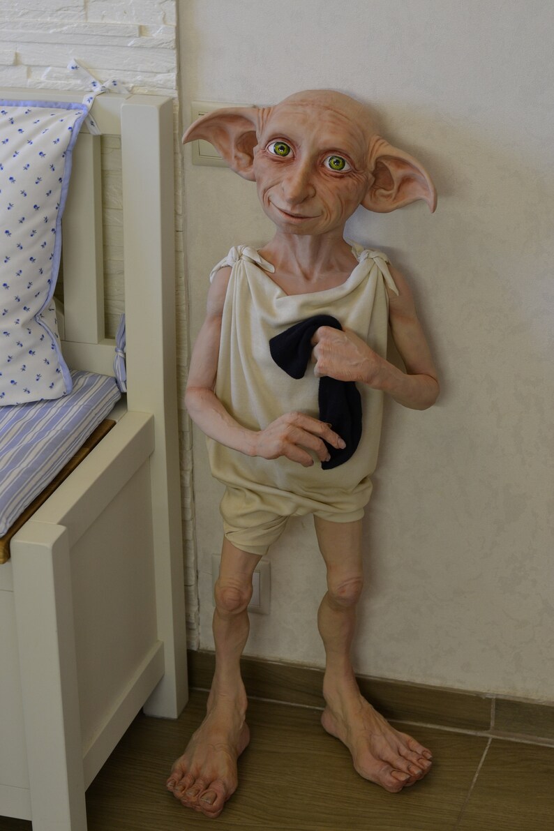 May include: A whimsical elf doll with large pointed ears, green eyes, and a light brown complexion. The doll is wearing a white tunic with a black sock tucked into the front. The doll is standing on a wooden floor.