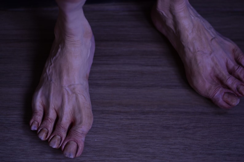 Puede incluir: Primer plano de dos pies descalzos con venas prominentes y dedos largos. Los pies descansan sobre una superficie de madera.