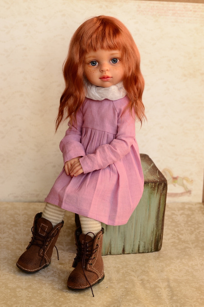 May include: A doll with long, wavy, red hair wearing a pink dress and brown boots. The doll is sitting on a wooden box.