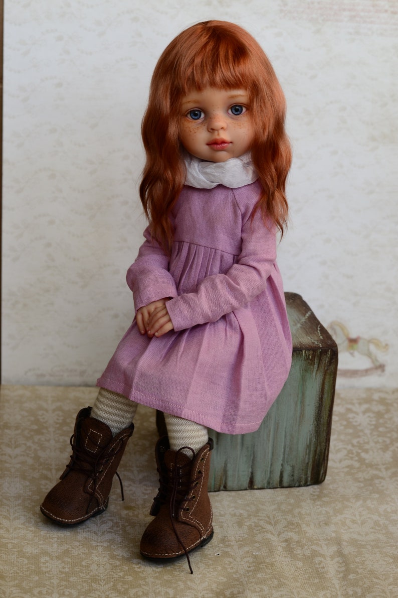 May include: A doll with long, wavy, red hair wearing a pink dress and brown boots. The doll is sitting on a wooden box.