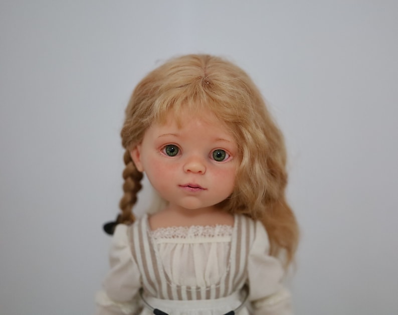 Puede incluir: Una mu&ntilde;eca de porcelana con el pelo rubio trenzado, que lleva un vestido a rayas blancas y marrones con un ribete de encaje.