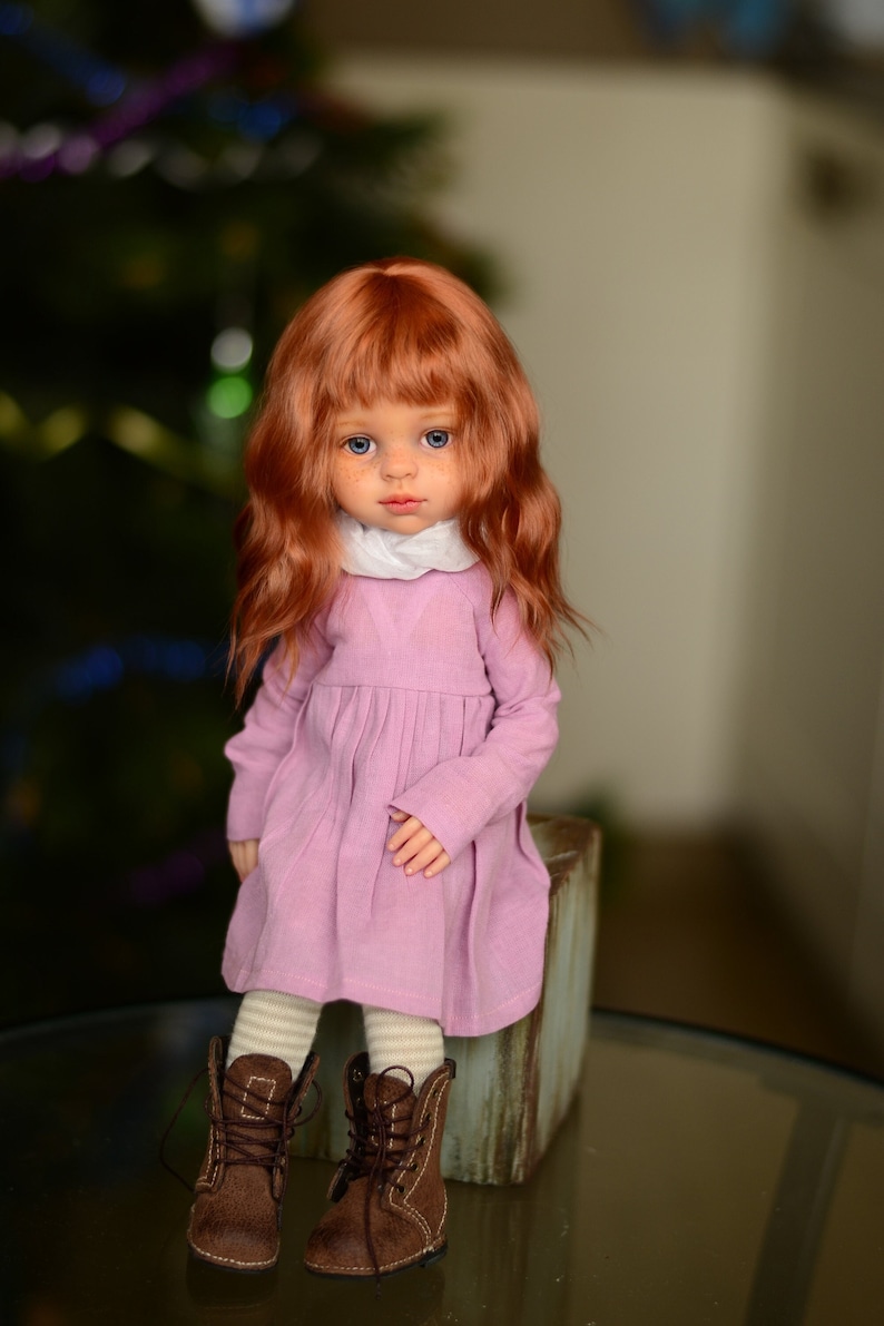 May include: A doll with long, wavy red hair wearing a pink dress and brown boots. The doll is sitting on a wooden block.