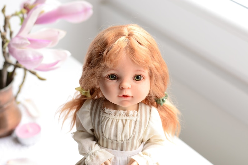 Puede incluir: Una mu&ntilde;eca de porcelana con el pelo largo, ondulado y casta&ntilde;o claro, que lleva un vestido a rayas blancas y marrones con un ribete de encaje. La mu&ntilde;eca tiene ojos verdes y una expresi&oacute;n dulce.