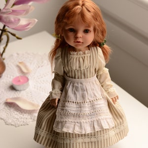 Puede incluir: Una mu&ntilde;eca de porcelana con el pelo rojo que lleva un vestido a rayas y un delantal blanco. La mu&ntilde;eca tiene ojos marrones y lleva zapatos marrones.