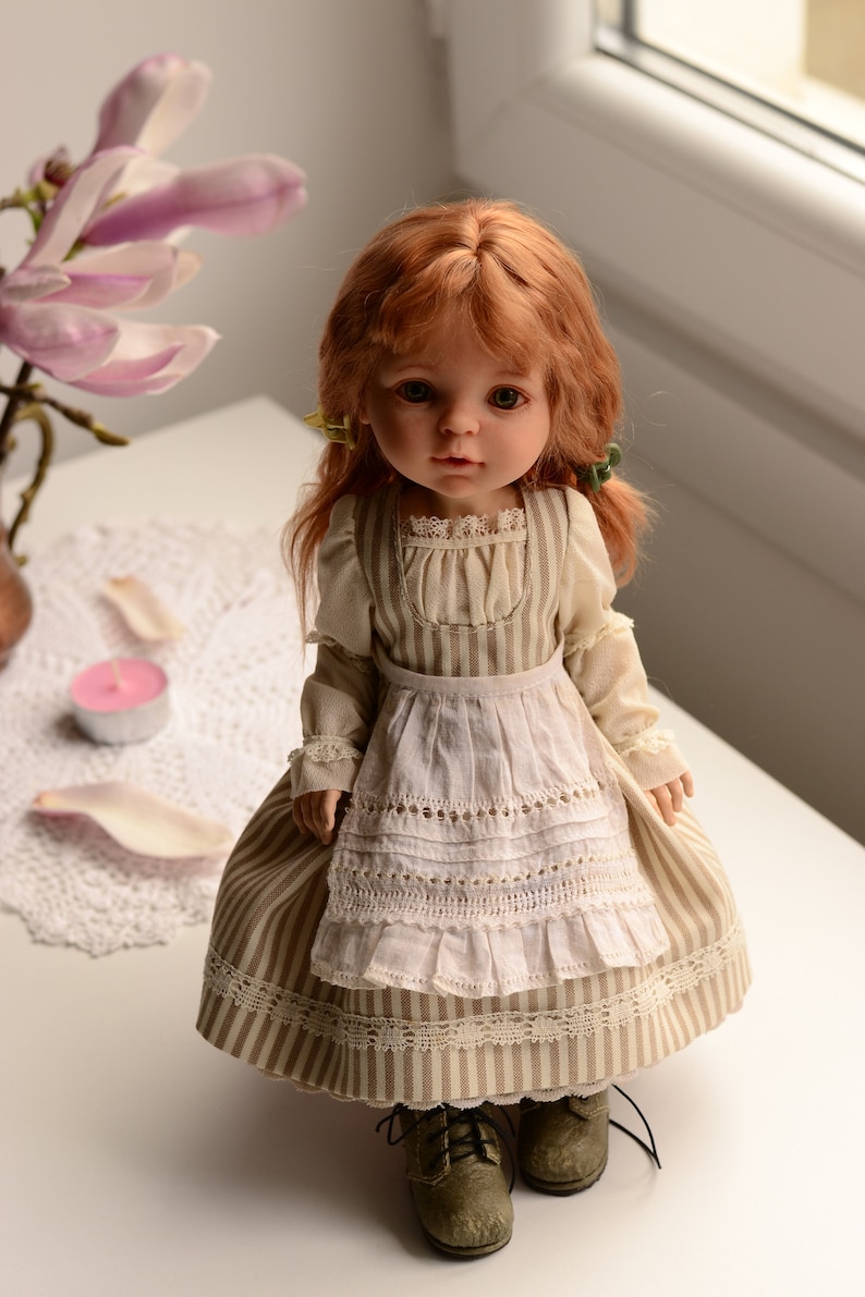 Puede incluir: Una mu&ntilde;eca de porcelana con el pelo rojo que lleva un vestido a rayas y un delantal blanco. La mu&ntilde;eca tiene ojos marrones y lleva zapatos marrones.