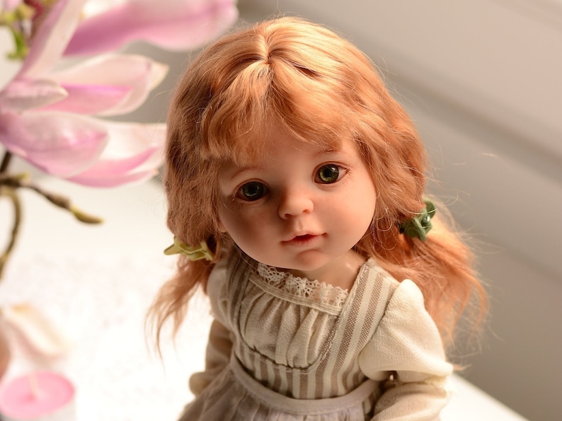 Puede incluir: Una mu&ntilde;eca de porcelana con el pelo largo, ondulado y casta&ntilde;o claro, que lleva un vestido a rayas blancas y marrones con un cuello de encaje. La mu&ntilde;eca tiene ojos verdes y una expresi&oacute;n dulce.