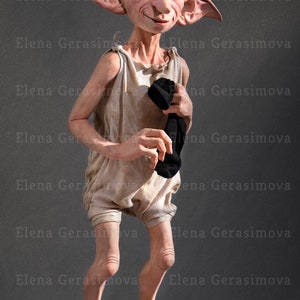 Puede incluir: Un primer plano de una mu&ntilde;eca de Dobby el elfo dom&eacute;stico. La mu&ntilde;eca lleva un mono blanco y sostiene un calcet&iacute;n negro. La mu&ntilde;eca est&aacute; de pie sobre un pedestal negro.
