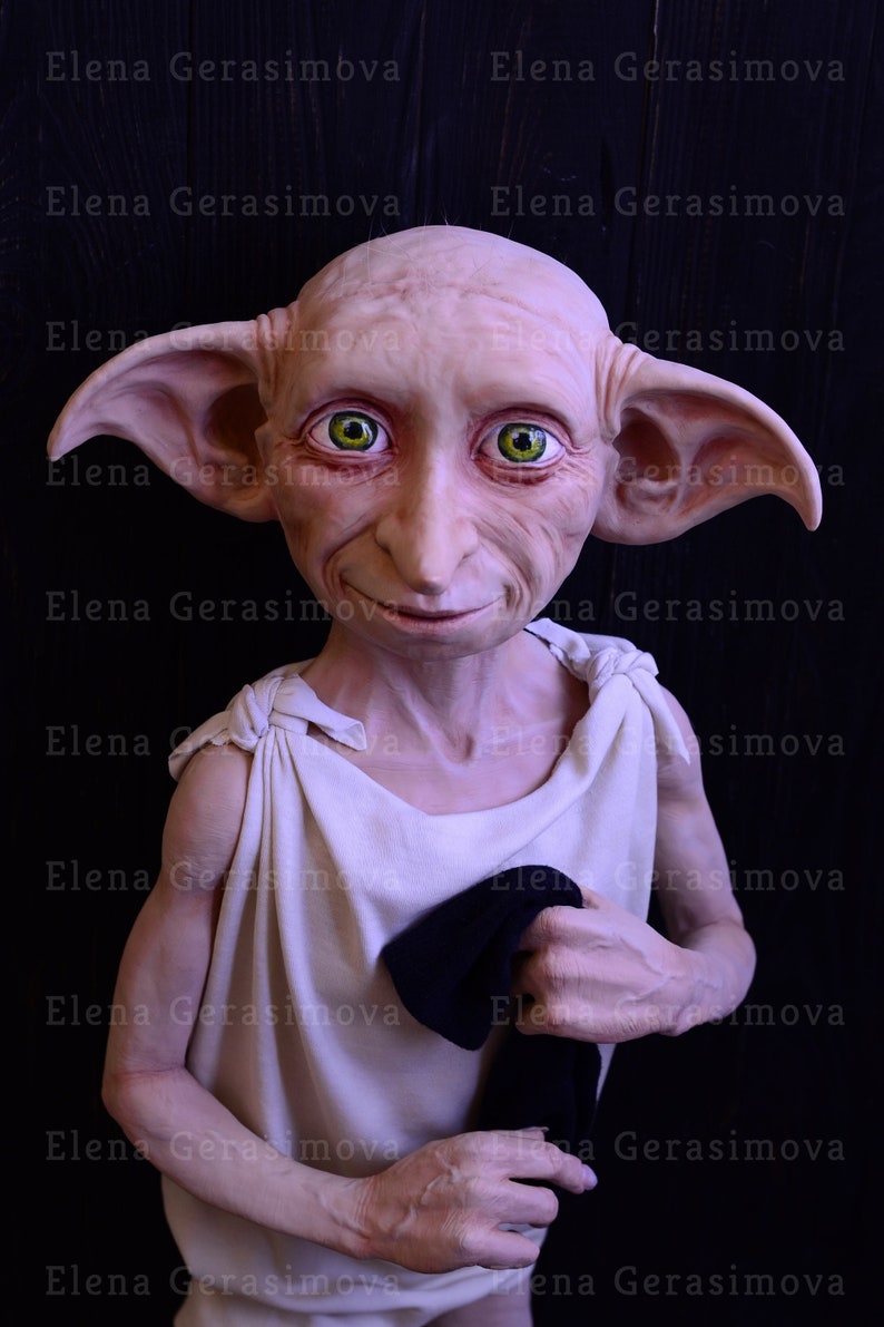 May include: A close-up of a Dobby the House-Elf doll. The doll is made of a light-coloured material and has large, pointed ears. The doll is wearing a white tunic and has a black cloth in its hand. The doll has green eyes and a friendly expression.