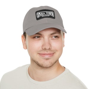 May include: A gray baseball cap with a curved brim. The cap features a black and white patch that reads "SMALL TOWN PROUD". The hat is made of a soft material and has a classic, casual style.