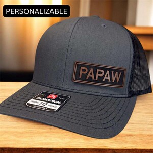 May include: A gray Richardson 112 trucker hat with a black mesh back and a brown leather patch that reads "PAPAW". The hat is adjustable and has a curved brim. The word "PERSONALIZABLE" is in the top left corner.