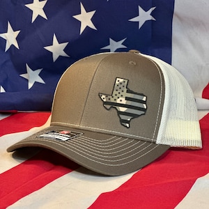 May include: A brown and white trucker hat with a black and white American flag Texas outline patch. The hat is on a red, white, and blue American flag.
