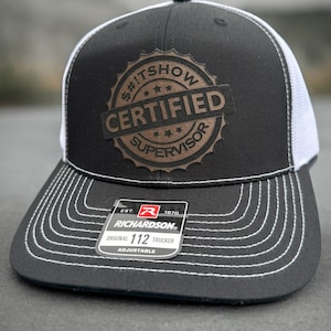 May include: Black and white trucker hat with a brown embroidered patch that says "$#!TSHOW CERTIFIED SUPERVISOR". The hat is adjustable and has a Richardson 112 label.