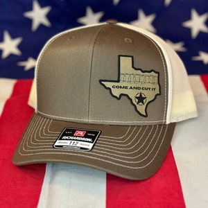Pode incluir: Um chapéu de caminhoneiro marrom e branco com um contorno dourado do estado do Texas com arame farpado ao redor da fronteira e o texto "Come and Cut It". O chapéu tem uma parte de trás de malha branca e uma parte frontal marrom com uma etiqueta de caminhoneiro Richardson 112.