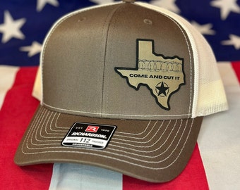 May include: A brown and white trucker hat with a gold outline of the state of Texas with barbed wire around the border and the text "Come and Cut It". The hat has a white mesh back and a brown front with a Richardson 112 trucker label.