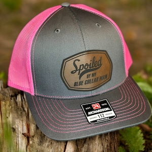 May include: A gray and pink trucker hat with a brown leather patch that says "Spoiled by my Blue Collar Man". The hat is adjustable and has a Richardson 112 label.