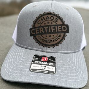 May include: Gray and white trucker hat with a brown leather patch that says "Chaos Certified Coordinator". The hat is adjustable and has a Richardson 112 label.
