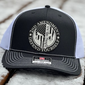 May include: Black and white trucker hat with a white mesh back. The front of the hat features a black and white patch with the text "2nd Amendment Defend Yourself" and a skull with an American flag design. The patch also includes the text "We the People".