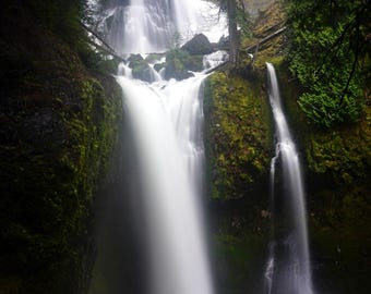May include: A scenic photograph of a waterfall cascading down a rocky cliff. The water is a blurred white, contrasting with the dark green moss and trees surrounding it. The image captures the natural beauty of a forest.