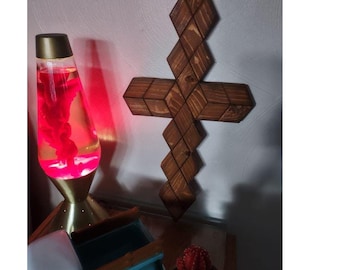May include: A wooden cross with a diamond pattern is mounted on a wall. The cross is made of stained wood. A lava lamp with red lava is to the left of the cross. A small blue truck and a cactus are on a shelf below.