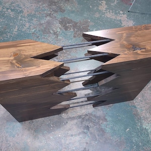 May include: A wooden console table featuring two dark brown stained wood blocks with jagged, interlocking edges connected by four horizontal grey steel rebar rods. The wood grain is visible on the top surfaces of the two main structural components.