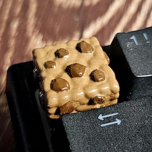 Puede incluir: Una tecla personalizada con forma de galleta marrón con chispas de chocolate más oscuras, colocada sobre un teclado negro. Un accesorio único para entusiastas de la informática.