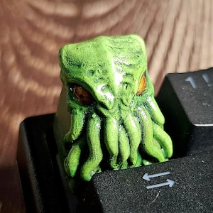 Puede incluir: Una tecla verde con temática de Cthulhu se encuentra sobre una tecla negra del teclado. La tecla presenta un diseño esculpido detallado con ojos naranjas y tentáculos. Las teclas del teclado son negras con letras blancas. Un accesorio único para teclados mecánicos.