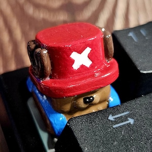 May include: A custom keycap depicting a cartoon character with a red hat and a white cross. The keycap is brown, blue, and red, designed for a keyboard. A unique accessory for computer enthusiasts. The keycap is a unique accessory for computer enthusiasts.