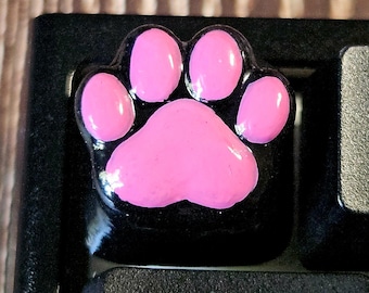 May include: A black mechanical keyboard keycap featuring a glossy, raised pink paw print design. The keycap is positioned on a keyboard next to a key labeled 1 and an F1 key, with a key below it marked with a double arrow and the word Macro.