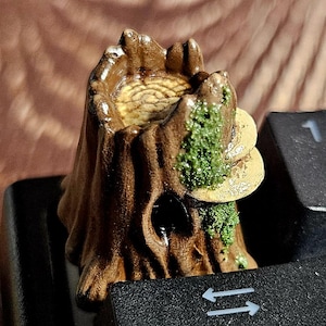 May include: A detailed tree stump keycap in brown and green, mounted on a black keyboard. The keycap is a custom design, with the "F1" and "Macro" keys visible. The keycap has a textured surface, adding to its unique appearance.