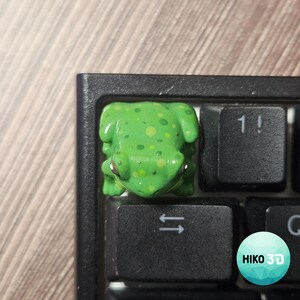 Puede incluir: Una tecla artesanal con forma de rana verde con manchas amarillas y verde claro, colocada en un teclado negro junto a una tecla con el s&iacute;mbolo 1! y una tecla con una flecha doble. Se observa un logotipo circular con el texto HIKO 3D.