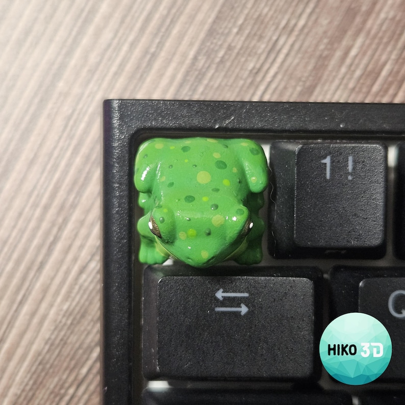 Puede incluir: Una tecla artesanal con forma de rana verde con manchas amarillas y verde claro, colocada en un teclado negro junto a una tecla con el s&iacute;mbolo 1! y una tecla con una flecha doble. Se observa un logotipo circular con el texto HIKO 3D.
