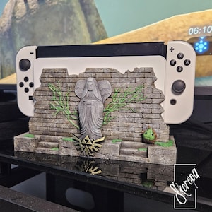 May include: A white Nintendo Switch console sits on a custom stand. The stand has a stone-like design with a statue, brick wall, and green details. A gold Triforce emblem is visible. The stand is designed to resemble a scene from the Legend of Zelda video game.