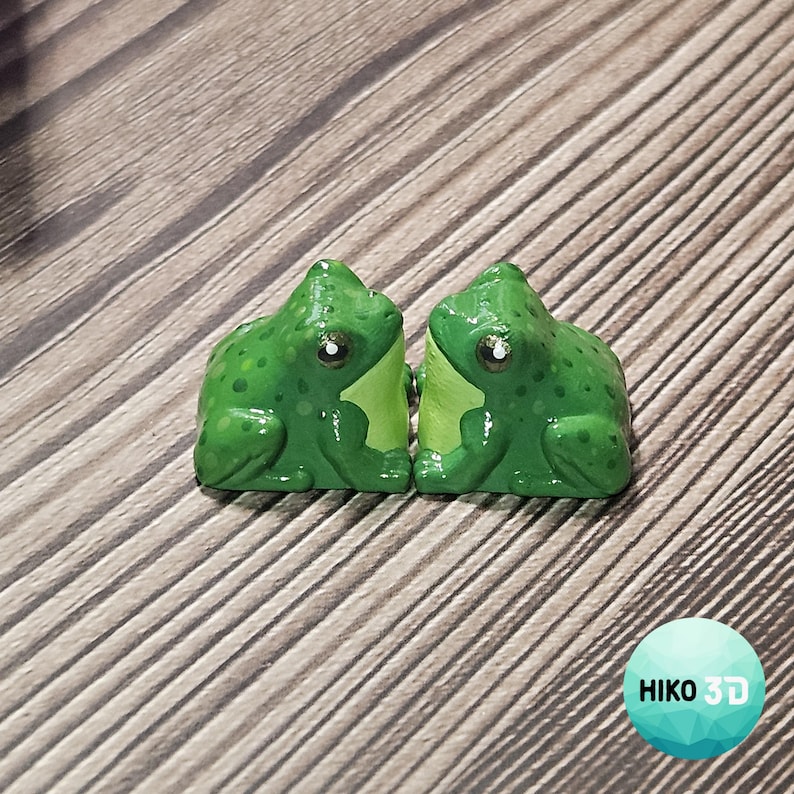Puede incluir: Dos peque&ntilde;as figuras de rana de color verde brillante con manchas verde oscuro y vientres de color verde claro. Cada rana tiene una forma lisa y redondeada con ojos visibles y una postura sentada. Las figuras est&aacute;n colocadas lado a lado.