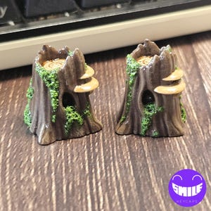 K&ouml;nnte beinhalten: Zwei braune Tastaturkappen in Baumstumpfoptik mit strukturierter Rinde, gr&uuml;nen Moosakzenten und hellbraunen Baumpilzen. Jeder Stumpf hat eine hohle &Ouml;ffnung mit dunklem Inneren und eine flache Oberseite, die die Maserung des Holzes zeigt.