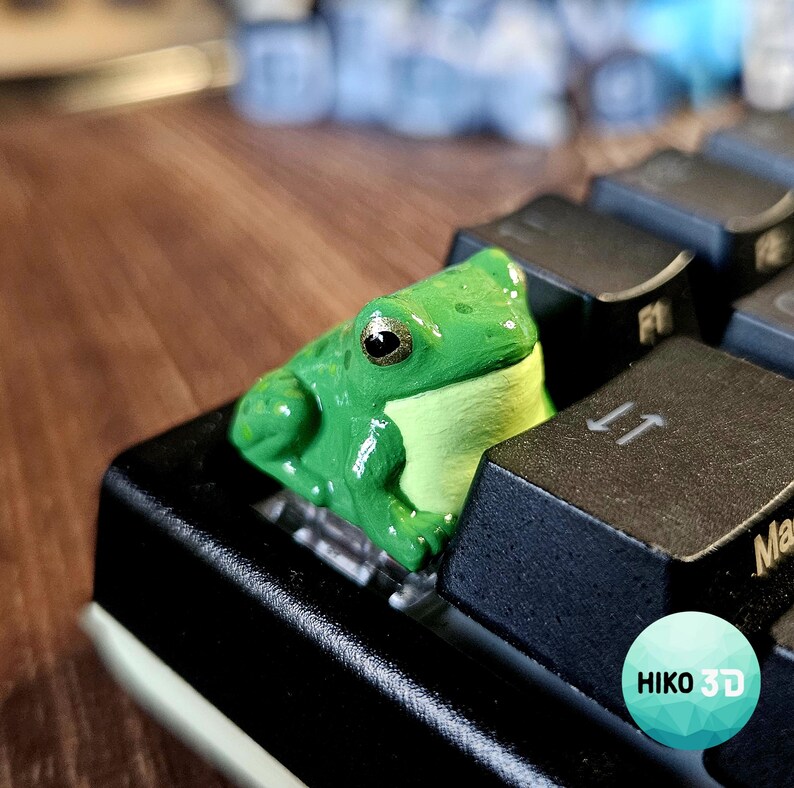 Puede incluir: Una tecla de teclado mec&aacute;nico con forma de rana verde, con el vientre amarillo claro y ojos negros, colocada en un teclado negro junto a una tecla con un s&iacute;mbolo de flecha bidireccional y el texto HIKO 3D en un logotipo circular.
