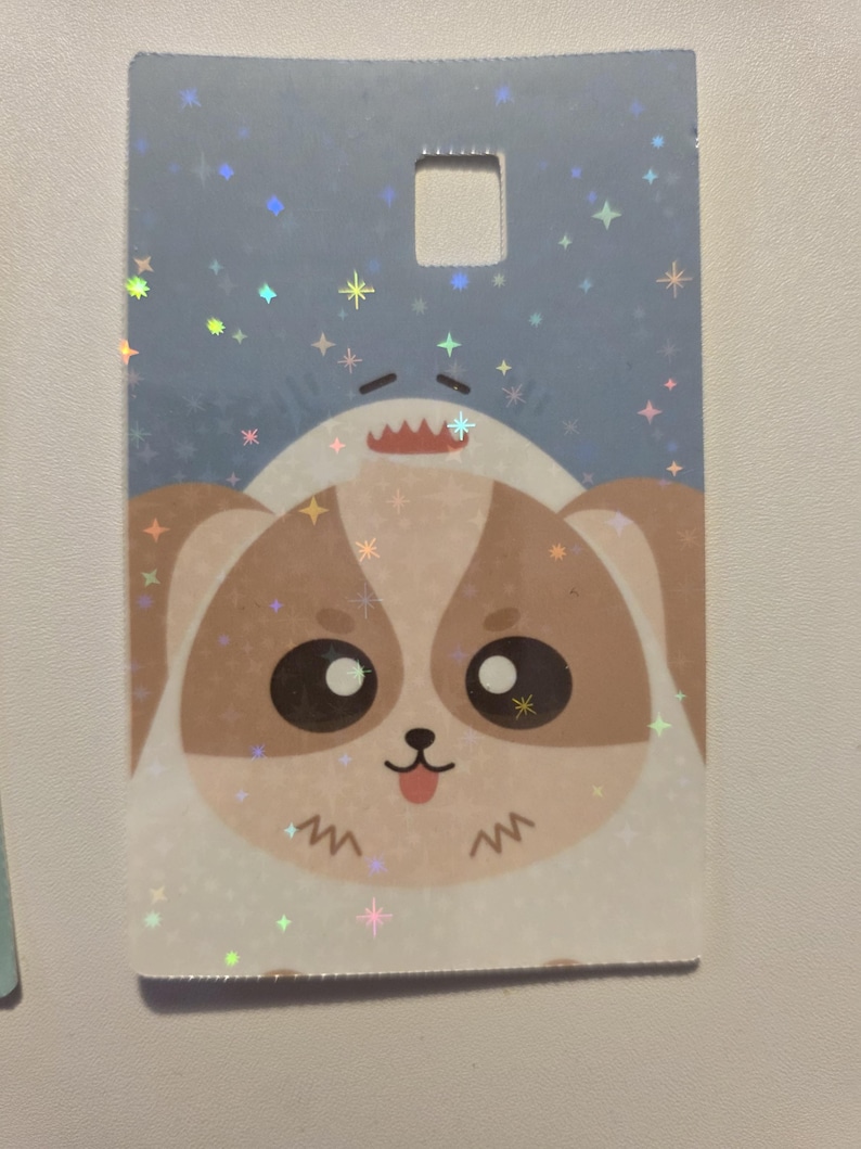May include: A rectangular tag with a cartoon dog and shark design. The dog has brown and white fur, large black eyes, and a pink tongue. The shark is white with a red mouth. The background is light blue with sparkling stars.