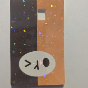 May include: A rectangular card with a split design: one half is dark brown, the other is orange. Both sides have a holographic star pattern. A white oval with a cartoon face is on the dark brown side.