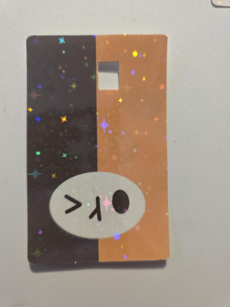May include: A rectangular card with a split design: one half is dark brown, the other is orange. Both sides have a holographic star pattern. A white oval with a cartoon face is on the dark brown side.