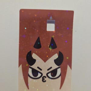 May include: A rectangular tag with a cartoon devil face. The tag has a brown background with a holographic glitter effect. The devil face has black horns, angry eyes, and a kissy face. There is a square hole at the top of the tag.