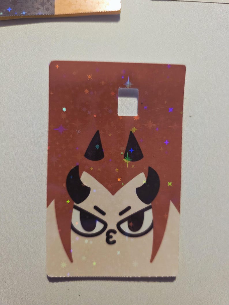 May include: A rectangular tag with a cartoon devil face. The tag has a brown background with a holographic glitter effect. The devil face has black horns, angry eyes, and a kissy face. There is a square hole at the top of the tag.