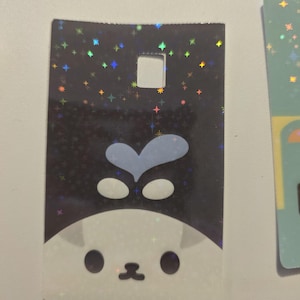 May include: A rectangular holographic card featuring a cartoon cat face with a blue heart-shaped design on top. The background is black with colorful star-shaped sparkles. The card has a square hole at the top.
