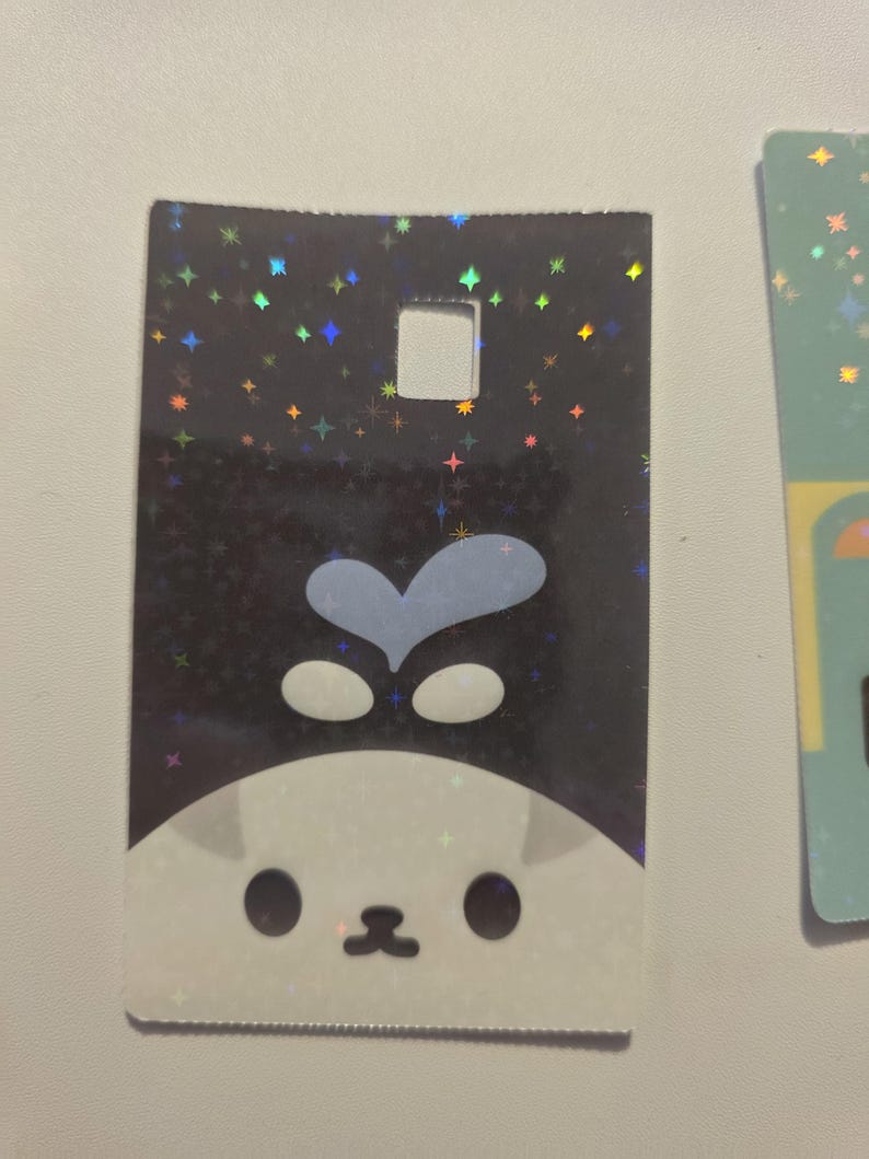 May include: A rectangular holographic card featuring a cartoon cat face with a blue heart-shaped design on top. The background is black with colorful star-shaped sparkles. The card has a square hole at the top.
