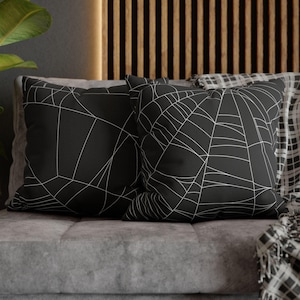 May include: Two black throw pillows with a white spiderweb design on a gray couch. A black and white plaid throw is draped over the couch.