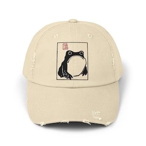 Könnte beinhalten: Eine beige, abgenutzte Baseballkappe mit einer Schwarzweißillustration eines Frosches in einem quadratischen Rahmen. Der Rahmen hat einen roten Stempel in der oberen linken Ecke.