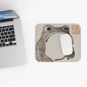 Puede incluir: Alfombrilla de ratón beige con una ilustración de rana marrón estilizada, con ojos grandes y boca curvada. Un ratón de ordenador blanco descansa sobre la alfombrilla. Un sello rojo está en la esquina superior derecha. La alfombrilla es rectangular.