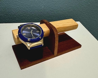 May include: A blue and gold wristwatch with a dark blue face rests on a wooden watch stand. The stand is made of light and dark brown wood, with a rectangular base and a curved support for the watch.