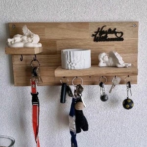 Könnte beinhalten: Holz-Schlüsselhalter mit einem Regal und Haken. Der Schlüsselhalter hat eine weiße Keramik-Engelfigur und ein Schild mit der Aufschrift "Herzlich Willkommen".