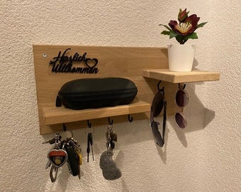 May include: Wooden wall-mounted key holder with a small shelf and hooks. The key holder has the words "Herzlich Willkommen" and a heart-shaped design. A black glasses case and keys hang from the hooks. A small white pot with a plant sits on the shelf.