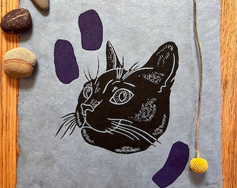 May include: A black and white linocut print of a cat's head with a purple background. The cat is looking to the right and has a surprised expression. The print is on a light blue paper.