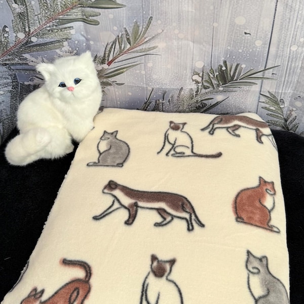 以下が含まれることがあります： 茶色、灰色、白の色合いで様々な猫のイラストが繰り返し描かれたクリーム色のフリースブランケットと、青い目とピンクの鼻を持つ小さなふわふわした白い子猫のぬいぐるみフィギュア。